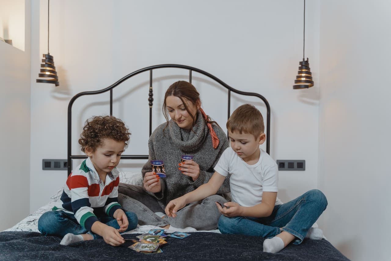 A mother and two young children playing an illustrated card game together on a bed