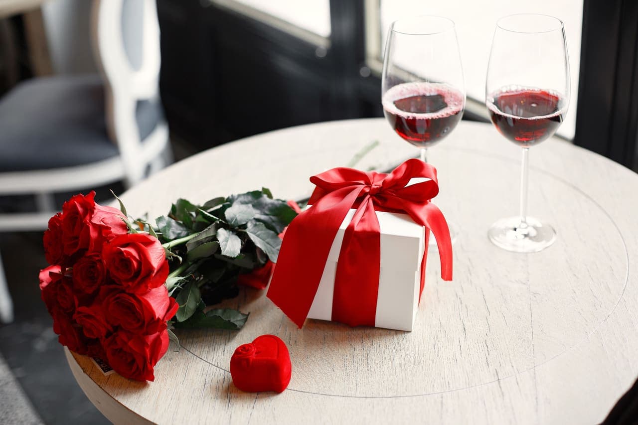 Red roses and a wrapped gift box beside two glasses of wine on a table