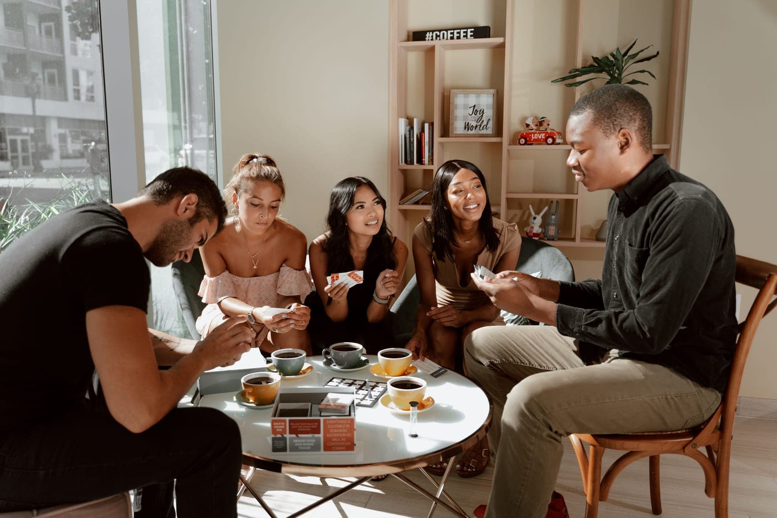 A group of friends around a wooden table mid-game, cards and wooden pieces spread between them in warm lamplight