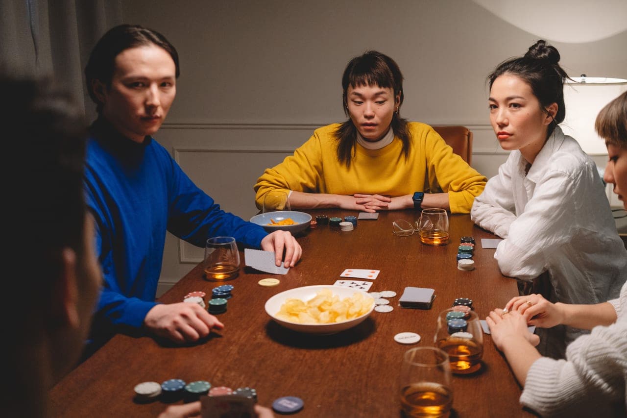 Six people gathered around a wooden table mid-game, cards and tokens spread between them